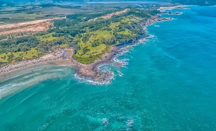Vista aérea de costão rochoso entre duas praias, com mar azul-turquesa e campos verdes