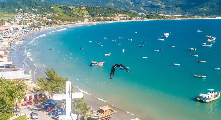 Enseada com barcos de pesca ancorados, praia, casario e uma cruz em primeiro plano, vista do alto