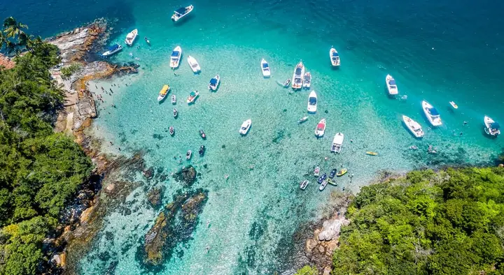 Lanchas e barcos em piscina natural de água cristalina entre costões cobertos de mata, vista de cima