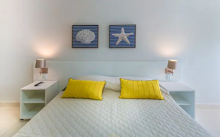 Quarto de casal com almofadas amarelas, arandelas e quadros de concha e estrela-do-mar sobre listras azuis