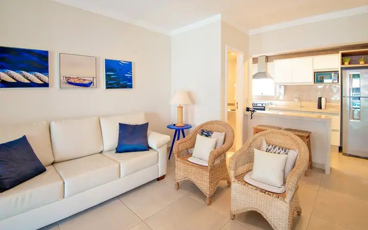 Sala de apartamento com sofá branco, almofadas azul-marinho, poltronas de fibra e quadros com fotos do mar