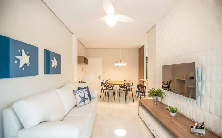 Sala de apartamento com sofá branco, quadros azuis de estrela-do-mar, TV em parede 3D e mesa de jantar ao fundo