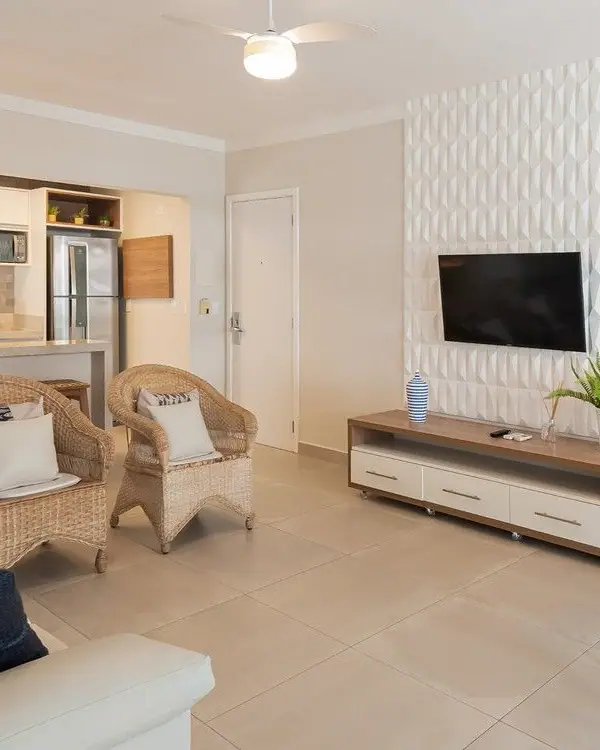 Sala integrada com TV em parede de revestimento 3D branco, poltronas de fibra, mesa redonda e cozinha ao fundo