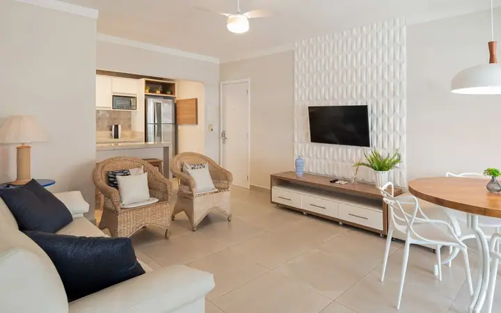 Sala integrada com TV em parede de revestimento 3D branco, poltronas de fibra, mesa redonda e cozinha ao fundo