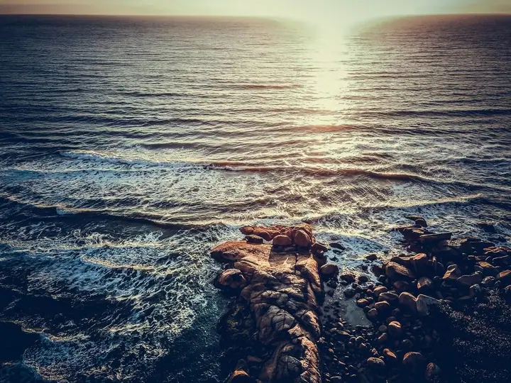 Brilho do sol na superfície do mar, com pedras escuras em primeiro plano