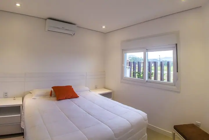 Quarto de casal claro com cama branca, almofada laranja, ar-condicionado e janela para o jardim
