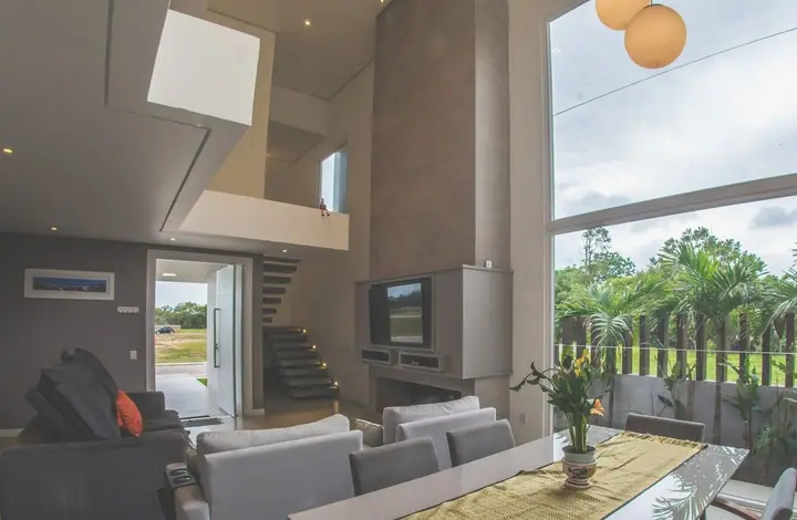 Sala de pé-direito alto com escada, sofás cinza, mesa posta e janelas de vidro voltadas para o jardim