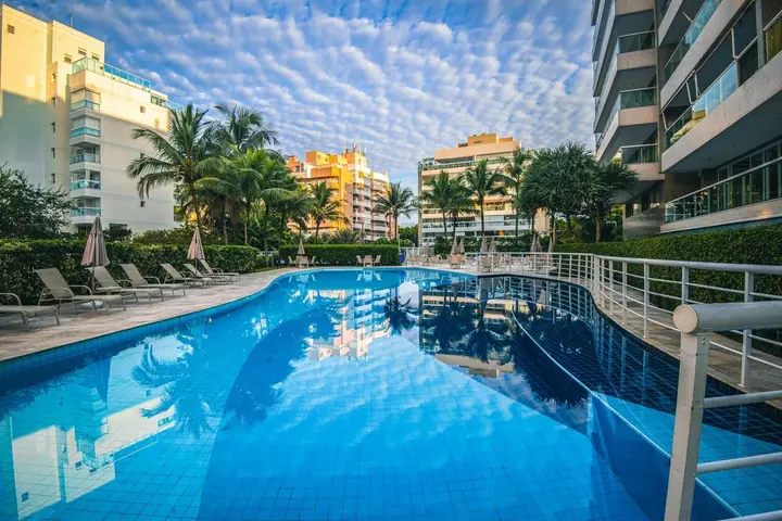 Piscina de condomínio com espreguiçadeiras, palmeiras e prédios ao redor sob céu com nuvens
