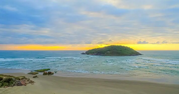 Ilha coberta de mata diante da praia, com o céu amarelado no horizonte