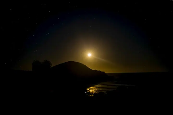 Lua nascendo atrás do morro e refletida no mar, em noite escura