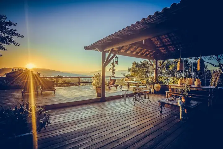 Deck de madeira com pergolado, mesas e luminárias ao sol baixo, com vista para o mar e os morros