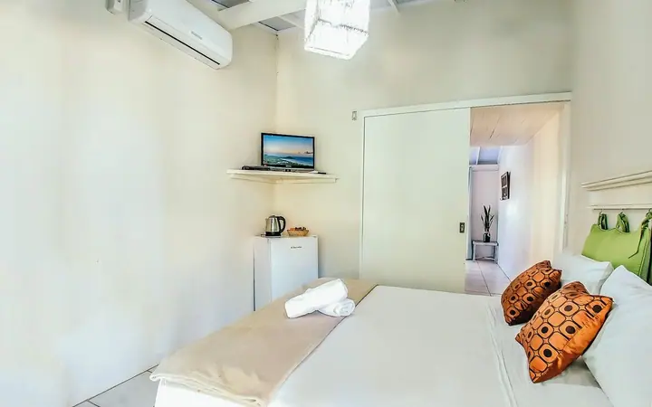 Quarto de pousada com cama de casal arrumada com enxoval branco, almofadas estampadas, toalhas, TV e frigobar
