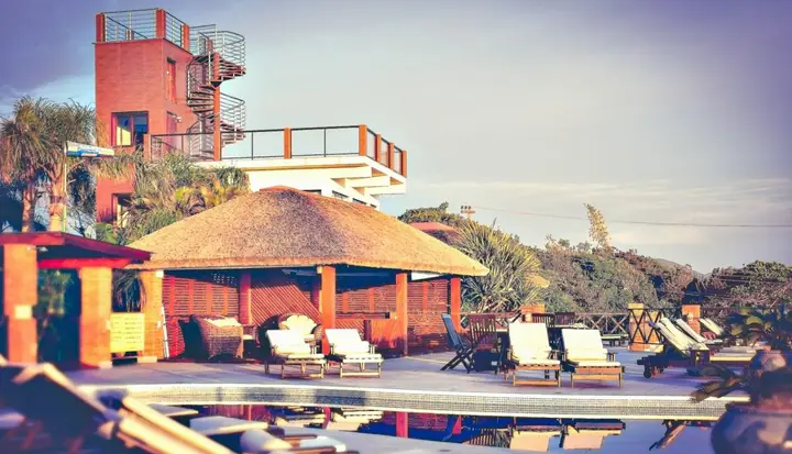 Área da piscina com quiosque de sapê, espreguiçadeiras e torre-mirante de tijolos entre a vegetação