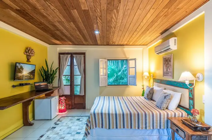 Suíte de pousada com forro de madeira, parede amarela, cama listrada, escrivaninha de madeira e porta para a varanda
