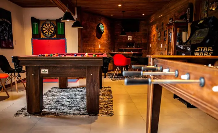 Sala de jogos com mesa de sinuca de feltro vermelho, pebolim, alvo de dardos, fliperama e cadeiras vermelhas