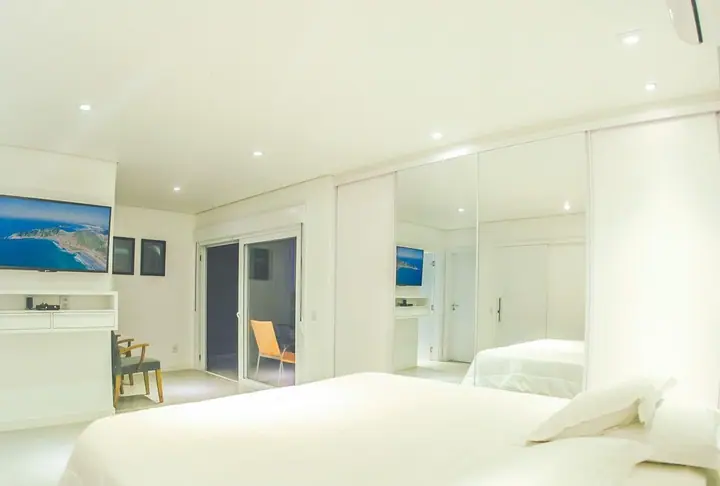 Suíte ampla e clara com cama de casal, armário espelhado, TV na parede e porta de vidro para a varanda