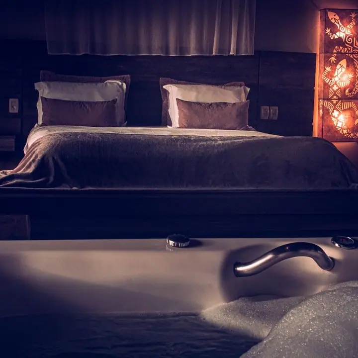 Suíte à meia-luz com banheira de hidromassagem em primeiro plano, cama de casal e luminárias de parede decoradas
