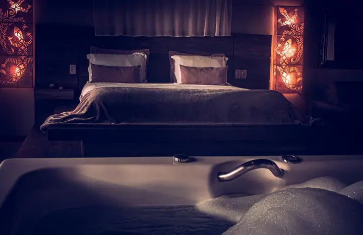 Suíte à meia-luz com banheira de hidromassagem em primeiro plano, cama de casal e luminárias de parede decoradas